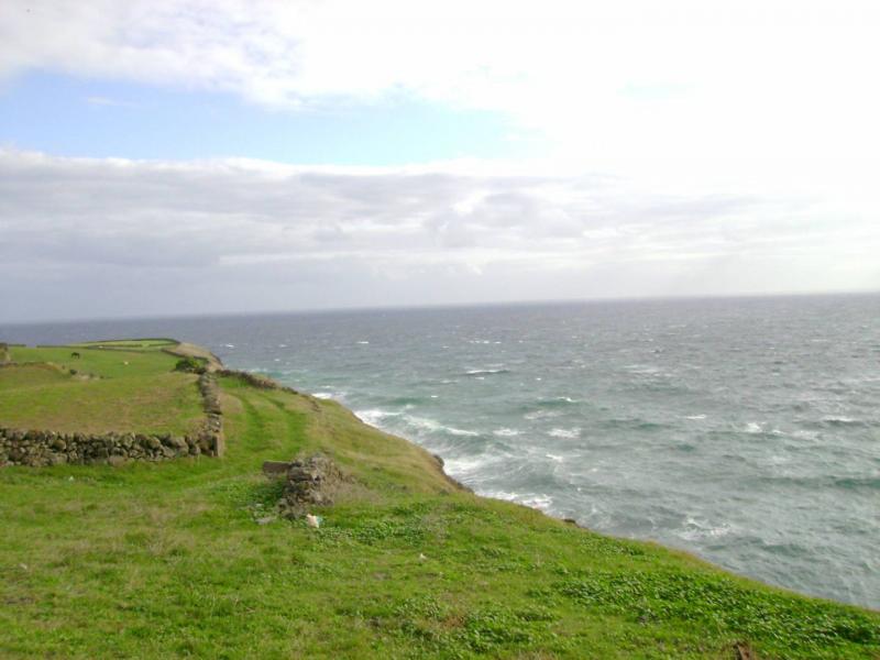 The images shows a spooky place in Portugal. It is a cliff in the Azores. We can see on the left green grass fields and on the right there is the endless ocean. On top the is the sky, cloudy, but with some sun.