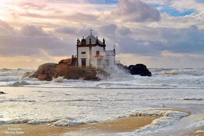 The picture shows an abandoned chapel on top of a rock, surrounded by the ocean. This is a spooky place in Portugal. The weather is very cloudy with some rays of sun coming out through the clouds. The sea ir very agitated.