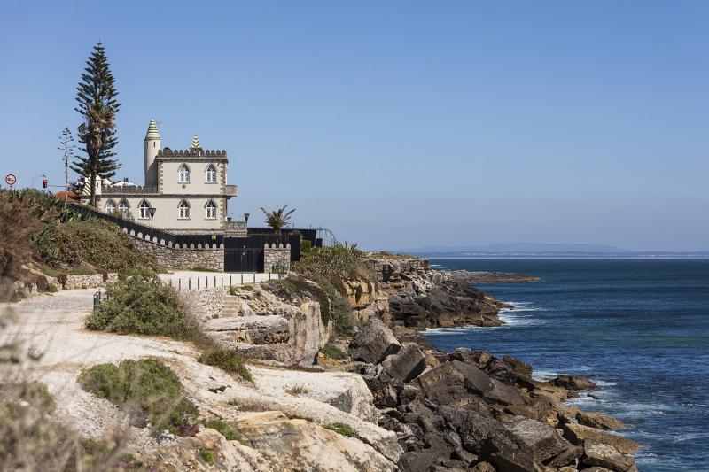 The picture shows a spooky place in Portugal. This one is a small castle in the coast of Estoril. On the left we see the building on the left speared by the sea through a sidewalk and rocks. The weather is a clear sky with a lot of sunlight.