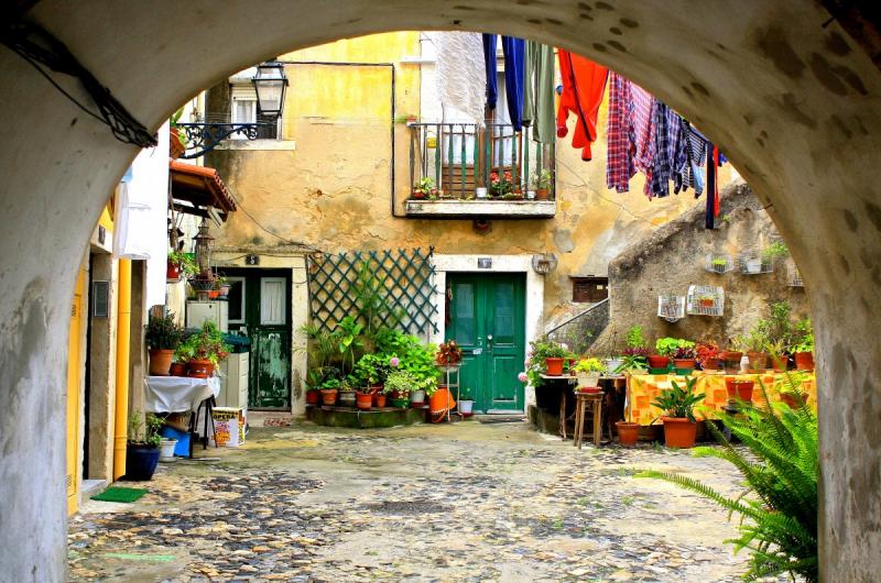 Spooky Place in Portugal, in Lisboa. The image shows a picture taken from the inside of a tunnel, rounded ceiling. In the end we see a typical Lisbon Patio, with a yellow building on the left, and a yellow building in the front, with two green doors. The colors are fading from age. There is a staircase on the façade on the left side. There are plant pots all around. There Is a balcony on the first floor and clothes hanging. 